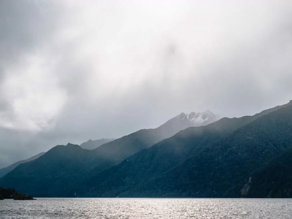 Sunlight pierces through dramatic clouds over the mountain-lined waters of Lake Manapouri, creating a soft glow on the lake’s surface