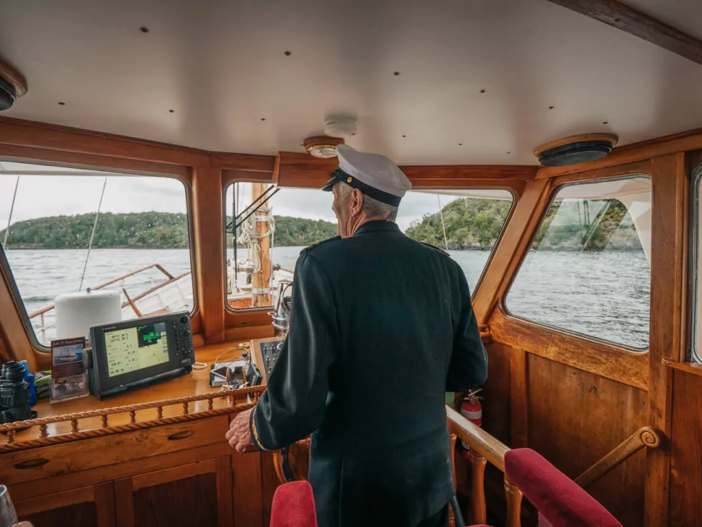 A ship captain steers a wooden boat from the helm, surrounded by old-fashioned instruments and large windows overlooking the water near Te Anau