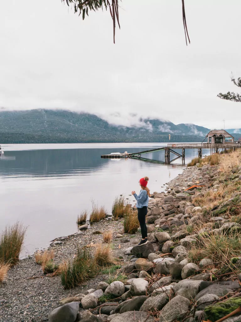 A woman in a red beanie looks out over the calm waters of Lake Te Anau from a rocky shoreline, with misty hills and a jetty in the distance