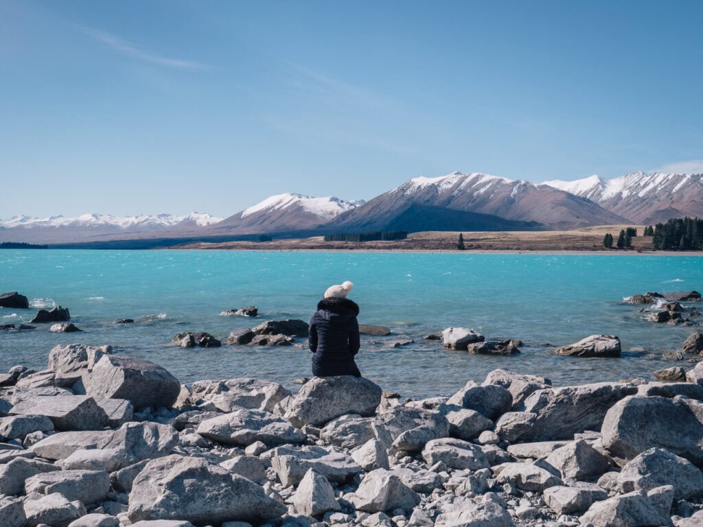Solo female traveller sitting on a rock in winter gear beside a snow-fringed Lake Tekapo on a clear blue day