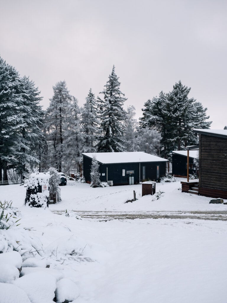 Small black cabin at Lakes Edge Holiday Park in Tekapo surrounded by snow and pine trees