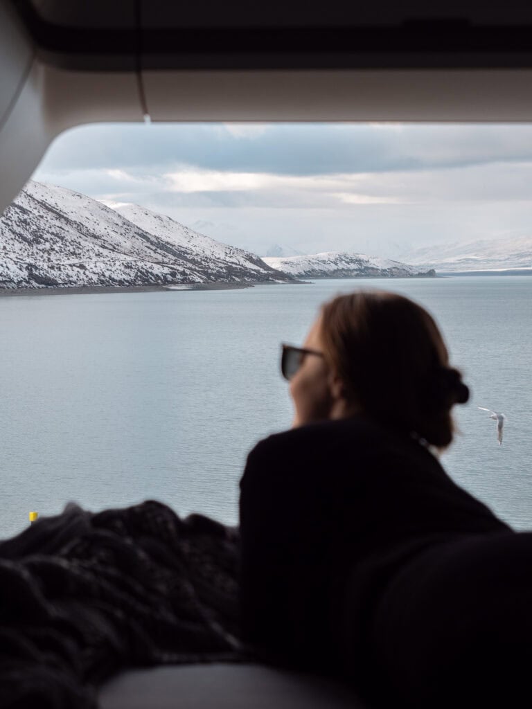 Alexx lying in the back of a campervan looking out across Lake Tekapo to snow-capped mountains