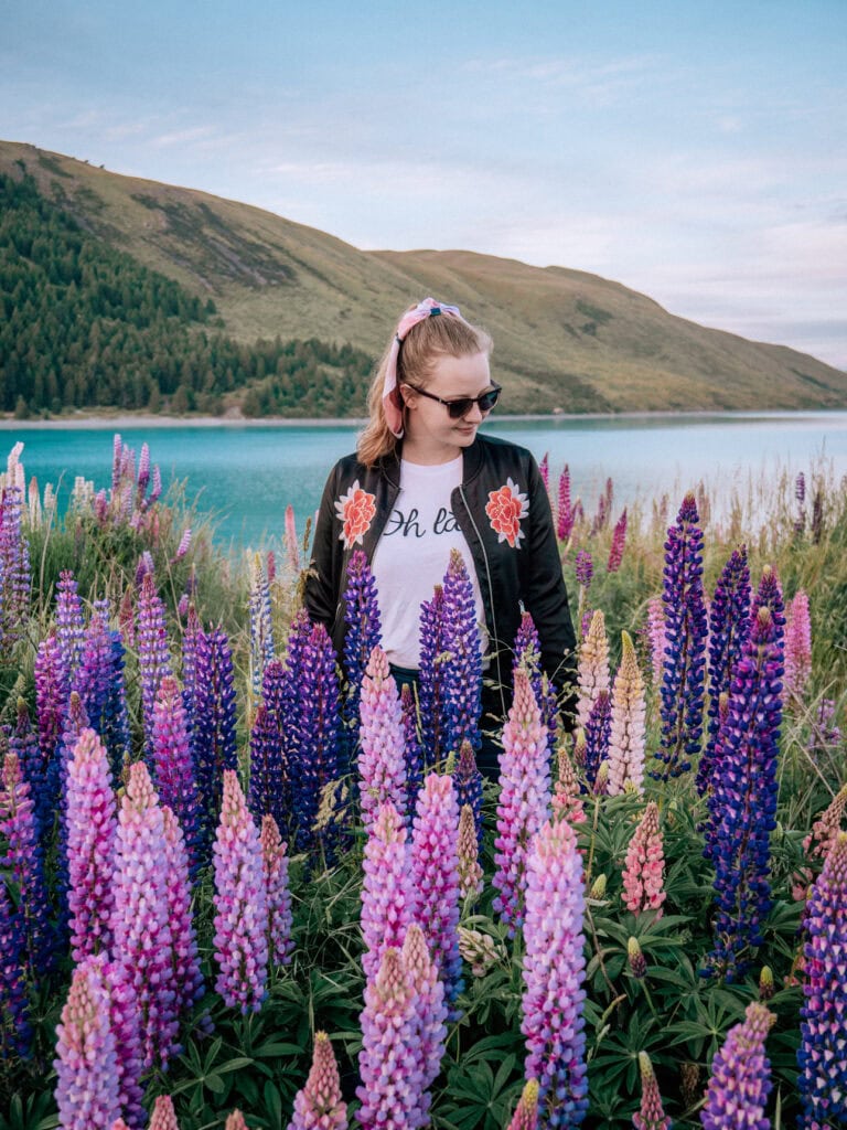 Alexx standing among tall pink and purple lupins on the shore of Lake Tekapo