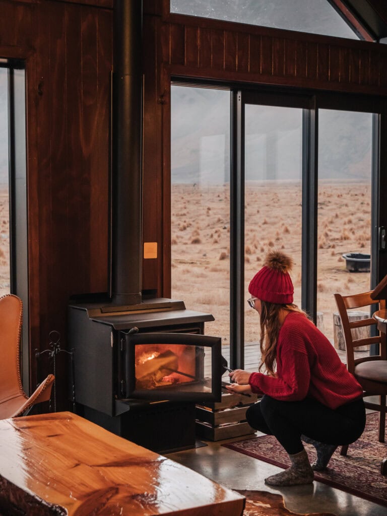 Alexx in a red beanie lighting the wood burner inside High Country Cabin in Twizel