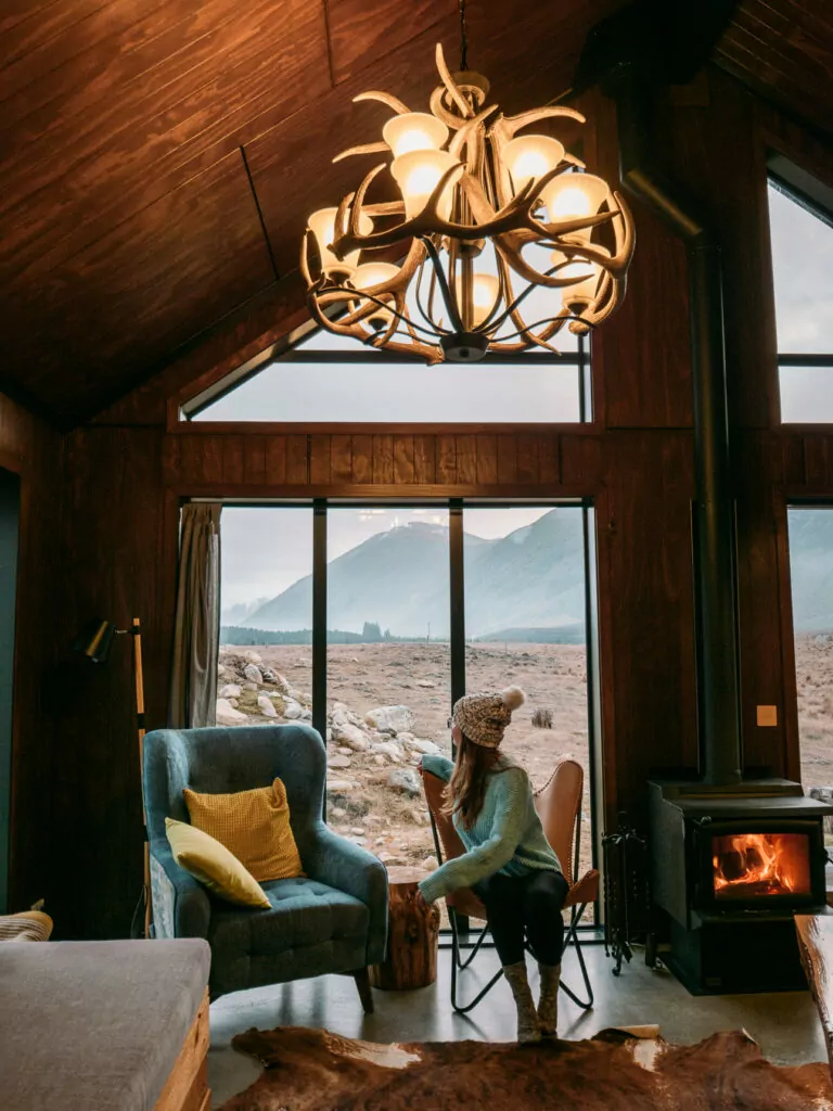 Cosy cabin interior with large windows framing golden tussock landscape views, featuring a woman sitting by a wood fire with geometric pendant light overhead near Twizel