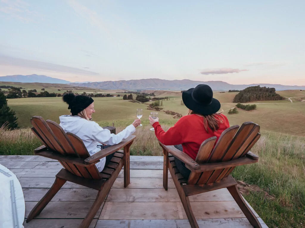 Two people drinking wine and enjoying the high-country landscape view from a remote Waitaki Valley deck at Valley Views glamping domes
