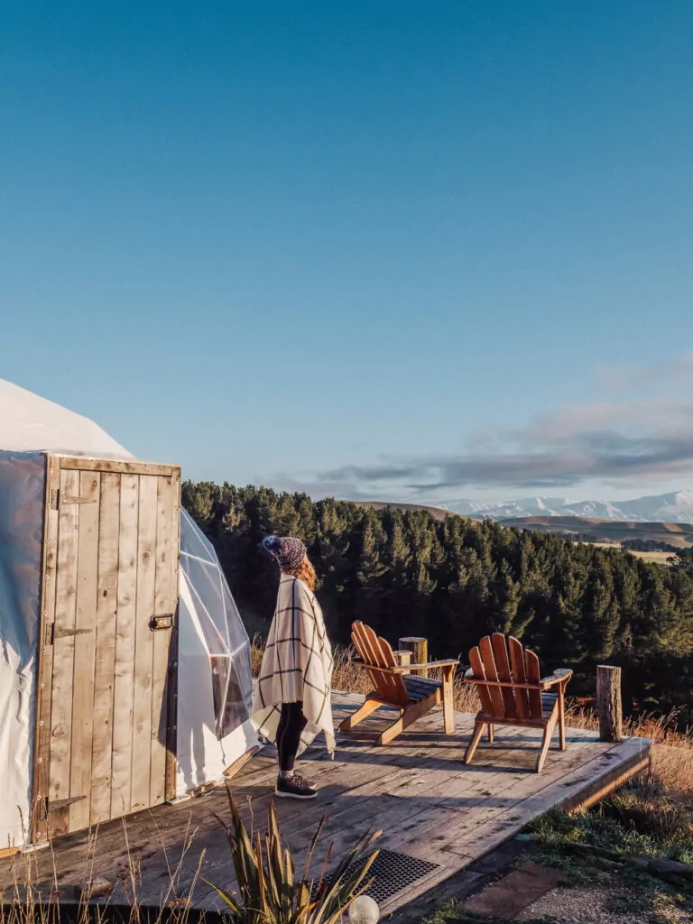 A person wrapped in a blanket stands outside a wooden yurt door, overlooking a scenic forested valley in New Zealand’s South Island. Two wooden chairs sit on the deck, facing the peaceful sunset view at Valley Views Glamping in Waitaki Valley