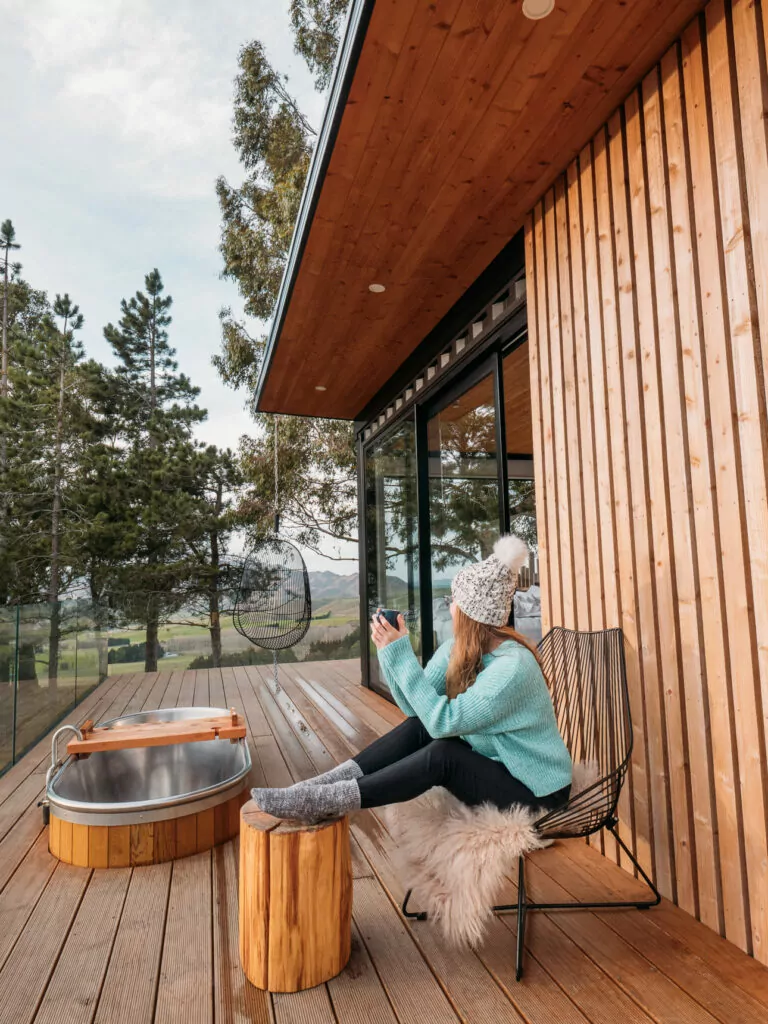 Woman relaxing in a turquoise jumper on a wooden deck with a cedar hot tub and forest views at Nest Treehouse glamping in Waitaki Valley