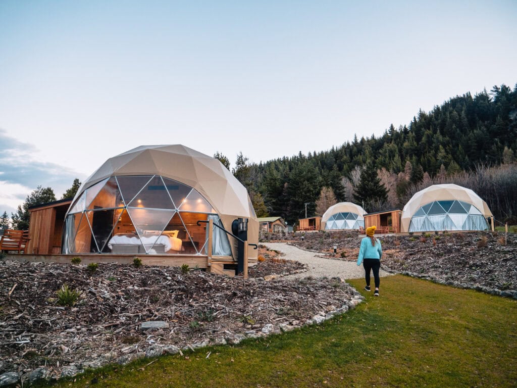 A woman walks toward glowing geodesic domes at Cross Hill Lodge in Lake Hāwea, set against a forested hillside at dusk