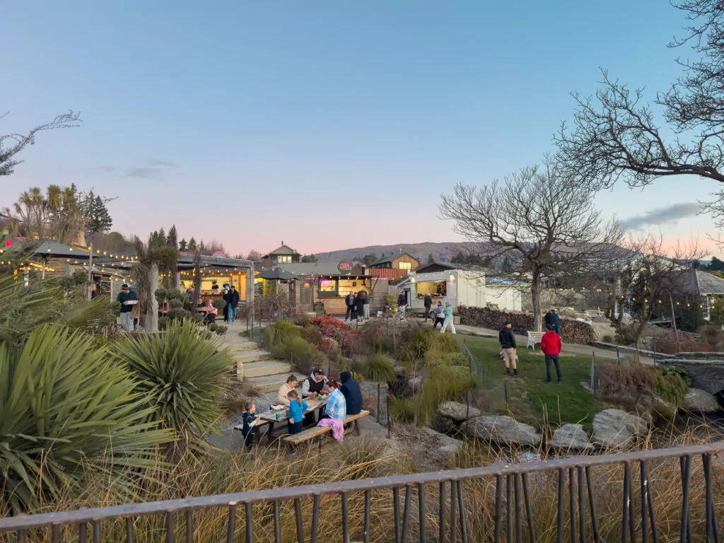 People relax in Wānaka at sunset, with string lights, rustic buildings, and surrounding mountain views