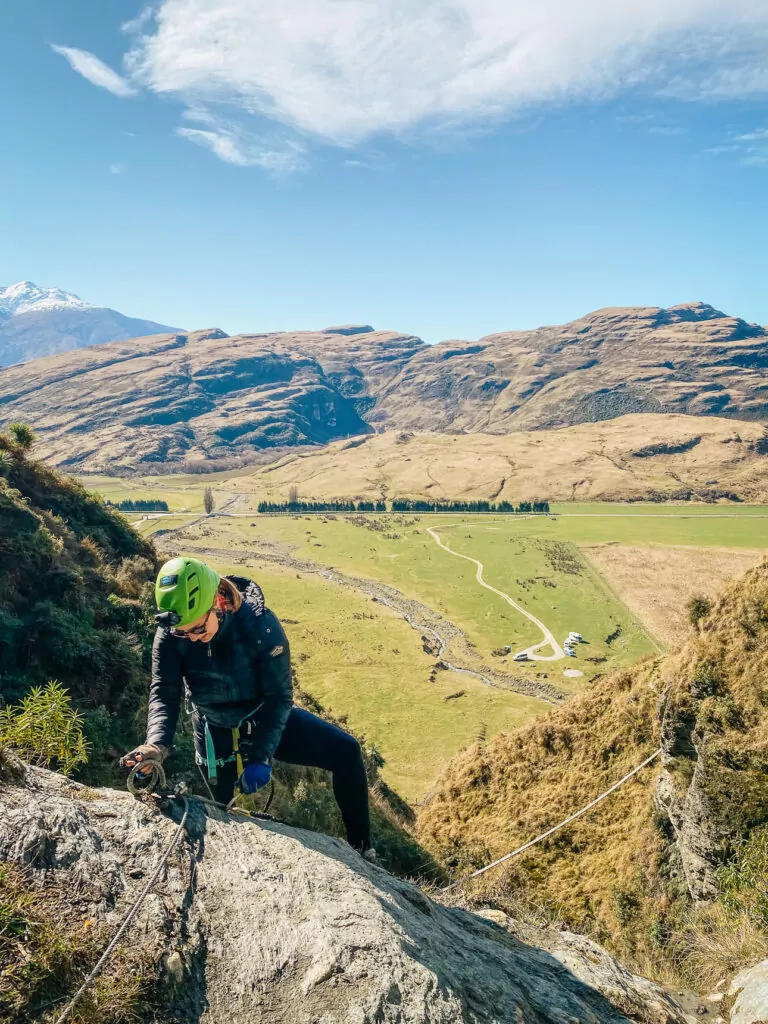 A climber scales a rocky outcrop near Wānaka on a via ferrata with Wildwire, with views of rolling farmland and mountains in the background on a clear day