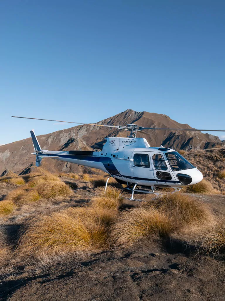A helicopter sits on a rugged mountain landing spot near Roys Peak in Wānaka, surrounded by golden tussock grass and alpine peaks