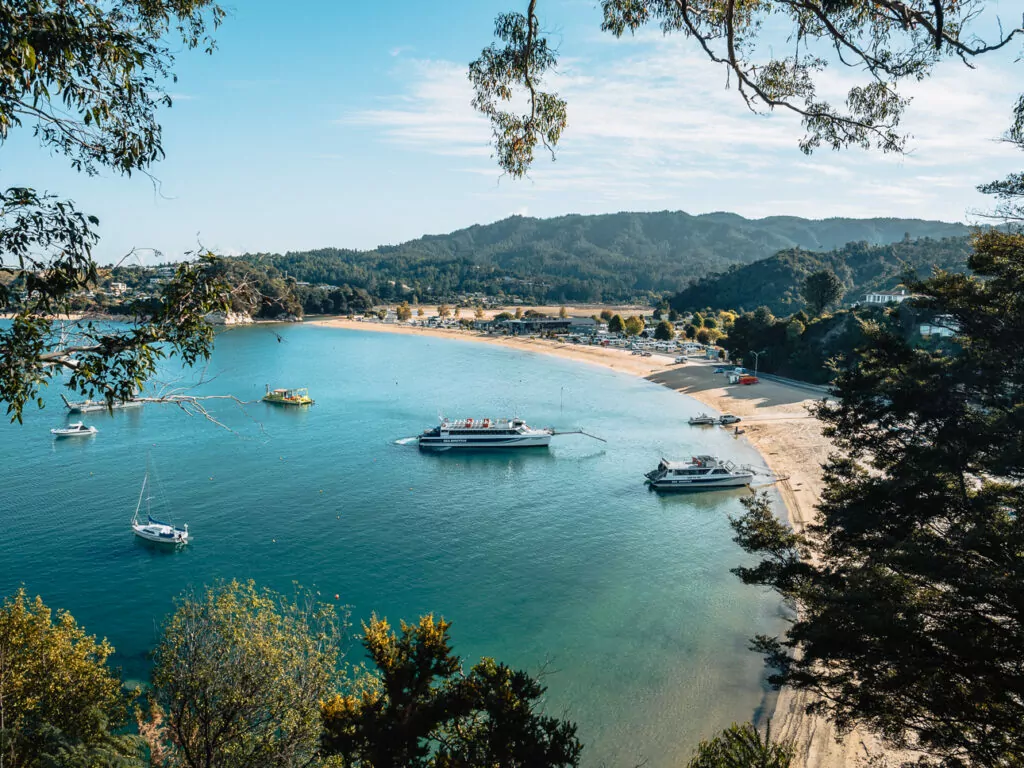 Golden sandy shoreline and clear turquoise water at Kaiteriteri Beach with anchored boats and forested hills