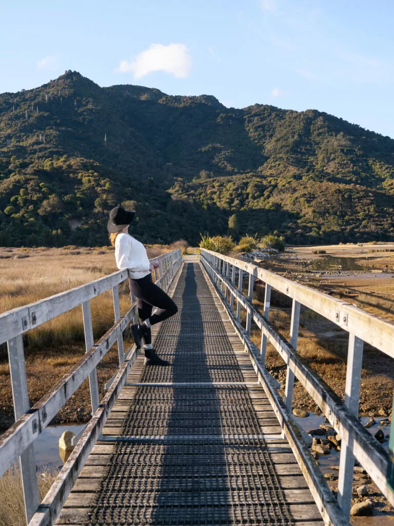 Hiker walking across a raised boardwalk near Marahau at the start of the Abel Tasman Coast Track, surrounded by open fields and hills