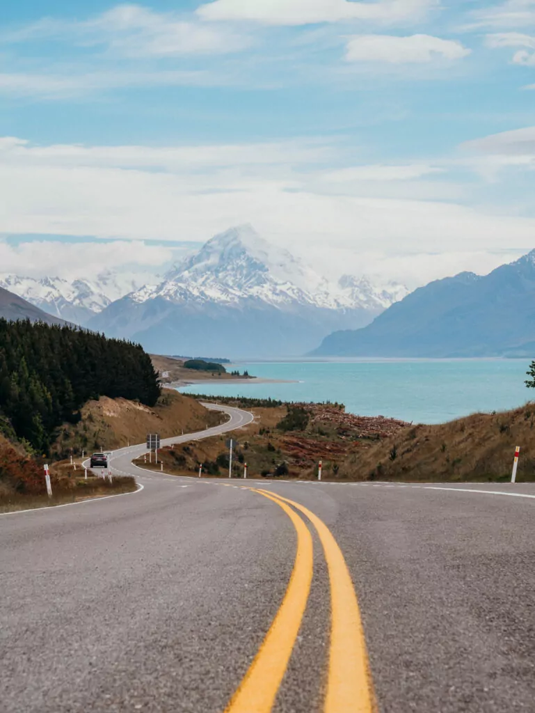 Curving road leading toward Lake Pukaki and Aoraki / Mount Cook framed by blue sky and snowy mountains