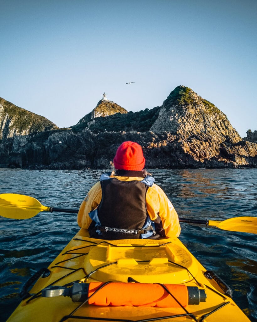 Kayaker paddling toward the rocky islets and lighthouse at Nugget Point in golden evening light