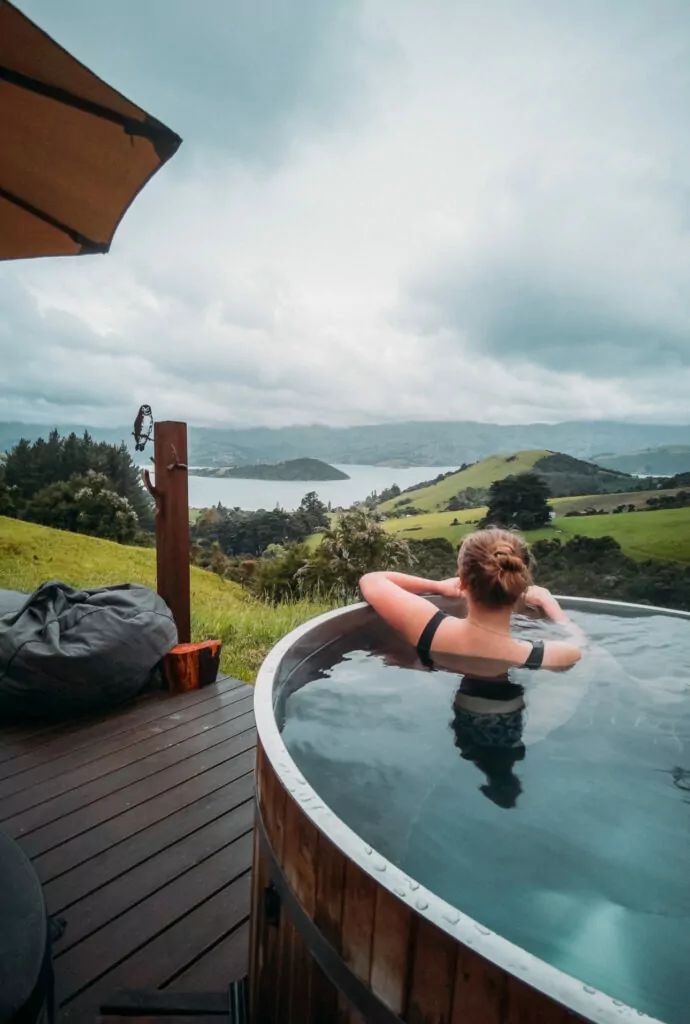 Woman soaking in a hot tub on the deck of Te Wepu pod with panoramic views of Banks Peninsula’s green hills and harbour.