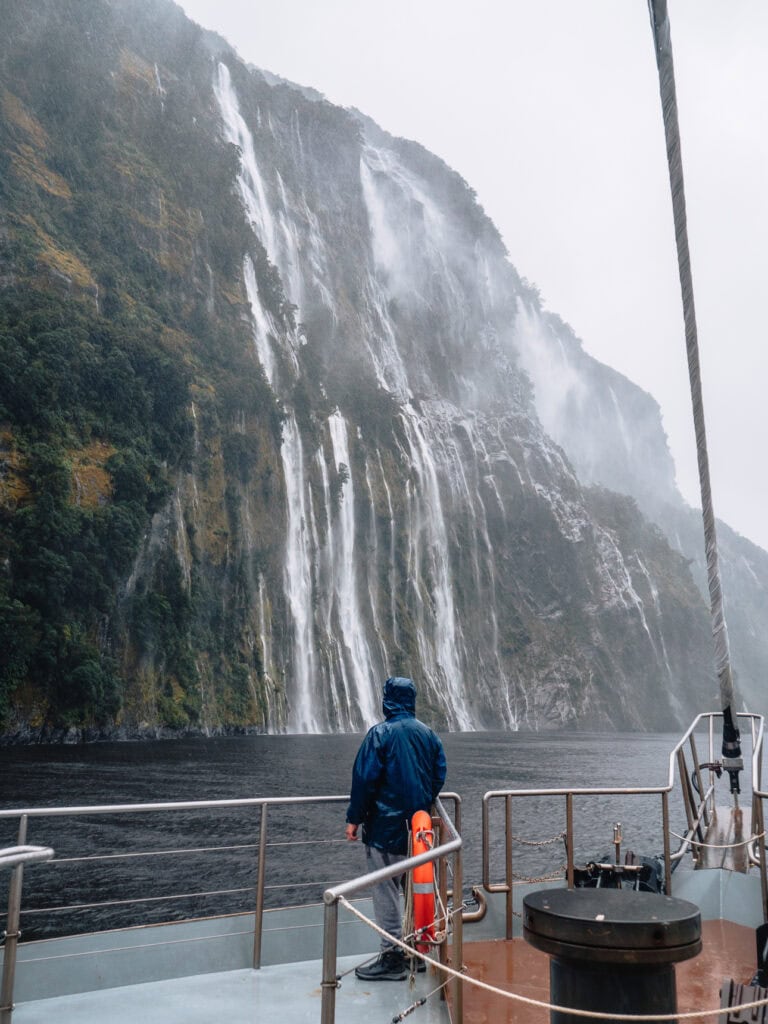 Visitor in a raincoat watching a tall waterfall cascade down misty cliffs into Doubtful Sound on a wet and moody day