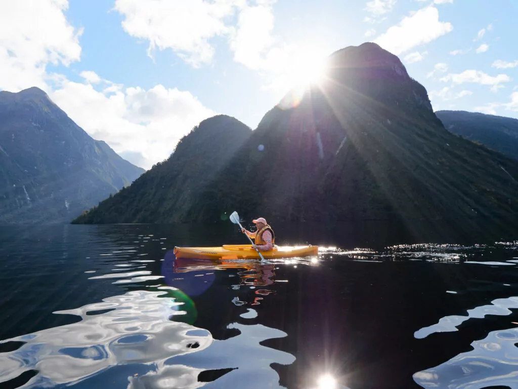 Solo kayaker paddling across glassy Doubtful Sound with the sun peeking over dramatic mountain silhouettes