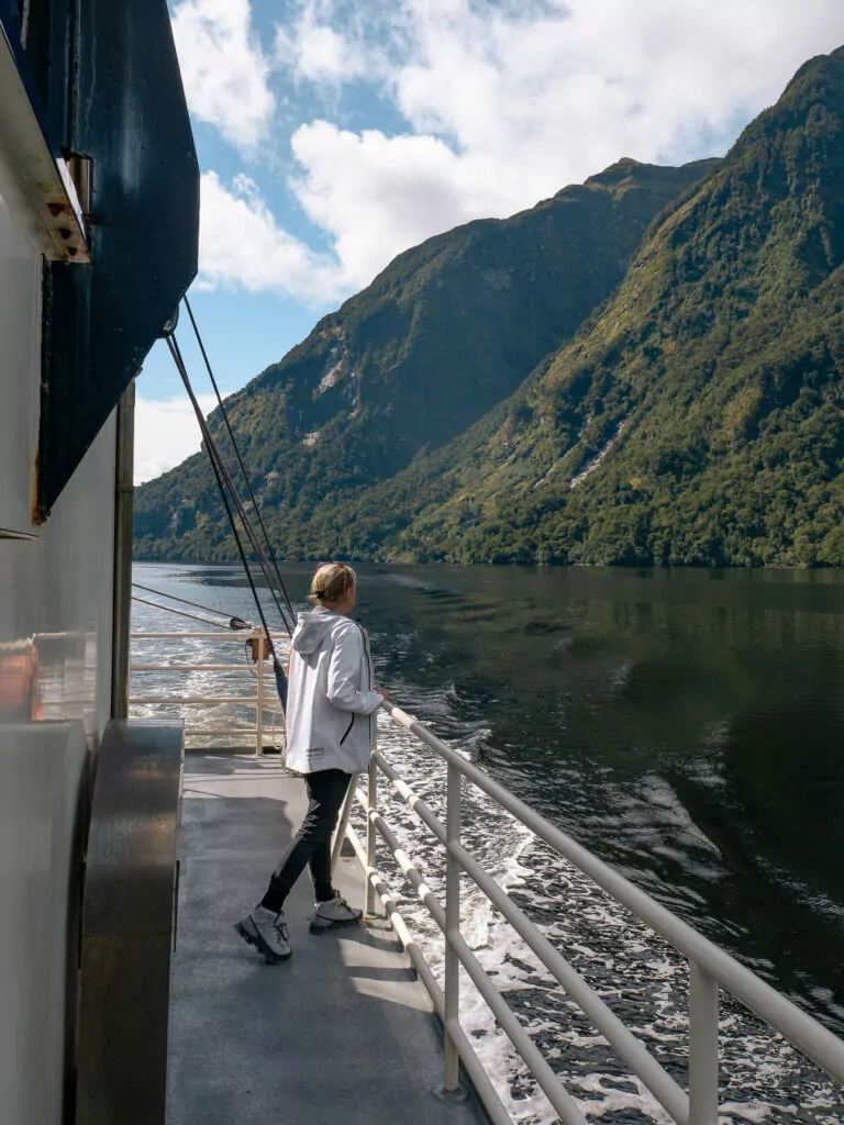 Traveller standing on the deck of a Doubtful Sound boat, looking out at steep fjord cliffs under blue skies