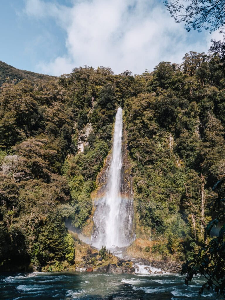 Tall, narrow waterfall plunging down a mossy rock face into a clear river pool surrounded by forest