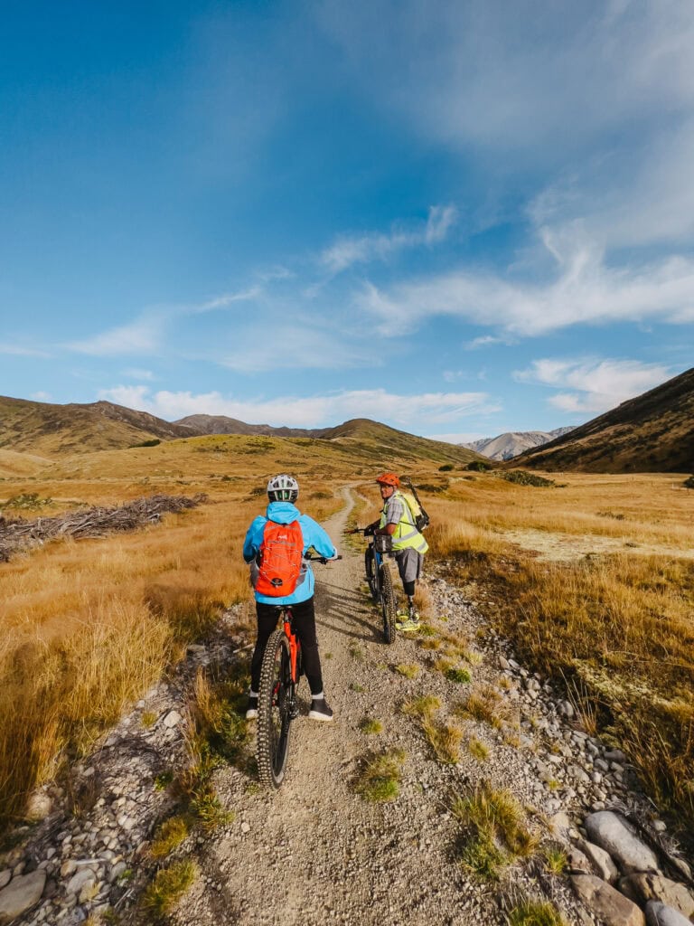 Two cyclists riding a gravel trail through golden tussock fields with mountain ranges in the distance.