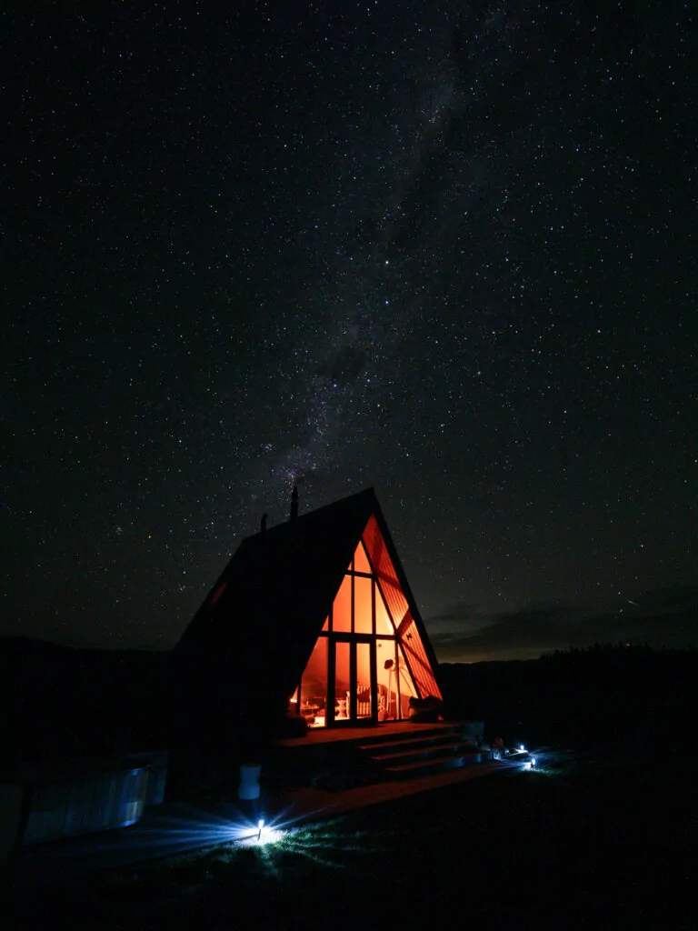 A-frame glamping cabin lit from within under a starry night sky in Hurunui on a South Island road trip itinerary