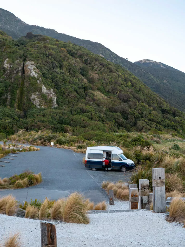 Campervan parked beside a still pond north of Kaikōura with mountain slopes and lush greenery reflecting in the water