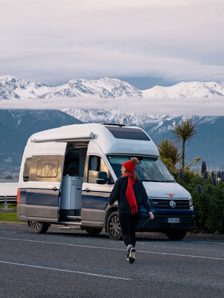 A woman in a red beanie walking towards a campervan parked along the Kaikoura coast with the snow-capped Seaward Kaikōura Range behind