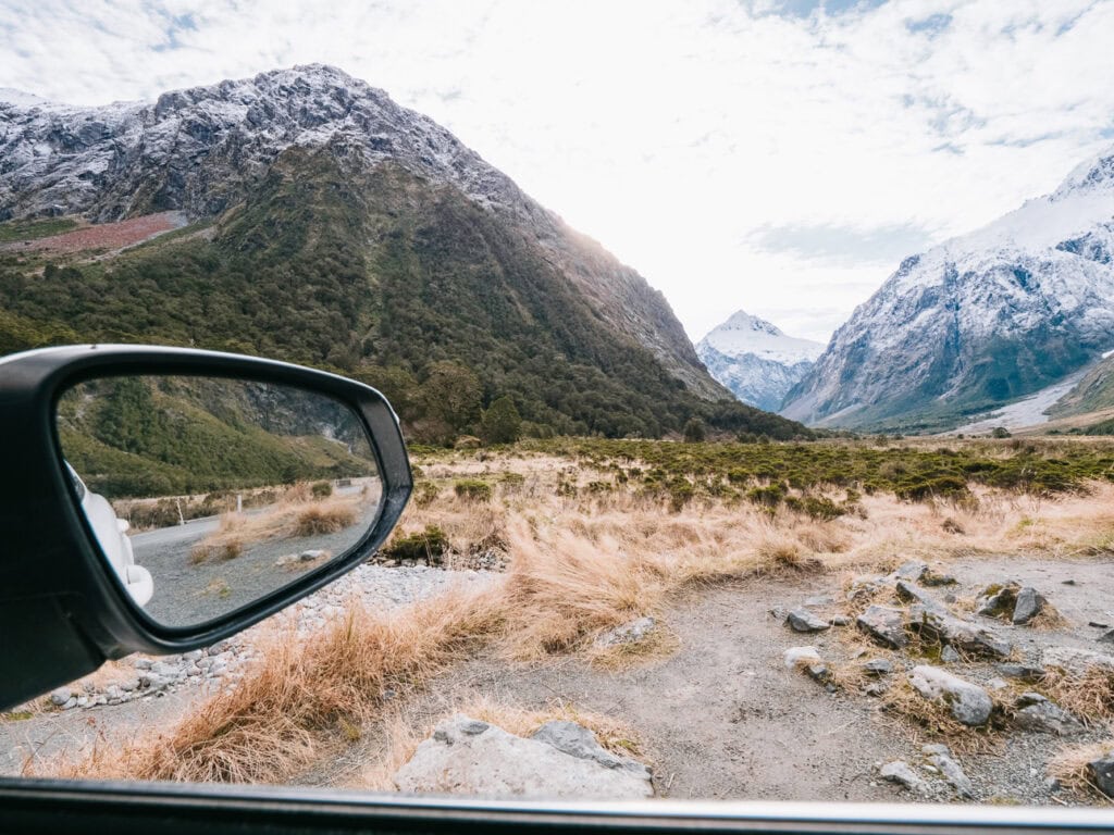 View through a car window of the Milford Road, with a grassy clearing and snow-covered mountains under an overcast sky
