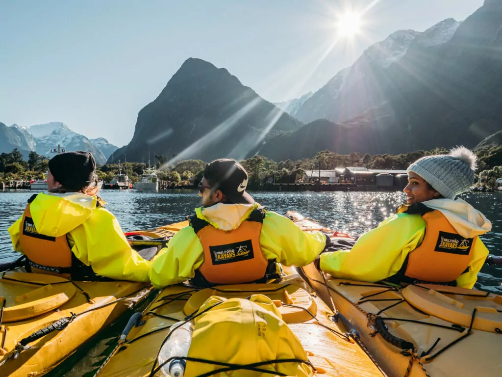 Group of kayakers in yellow jackets paddling into the sun on calm Milford Sound waters, surrounded by mountains