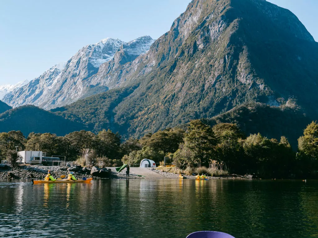 Low buildings and boats at the water’s edge with sheer peaks rising behind at the Milford Sound waterfront