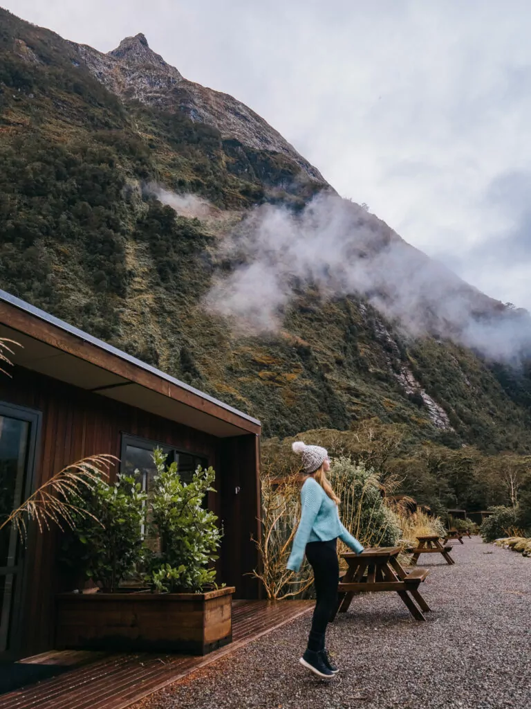 Traveller walking past wood cabins at Milford Sound Lodge with towering cliffs and mist in the background