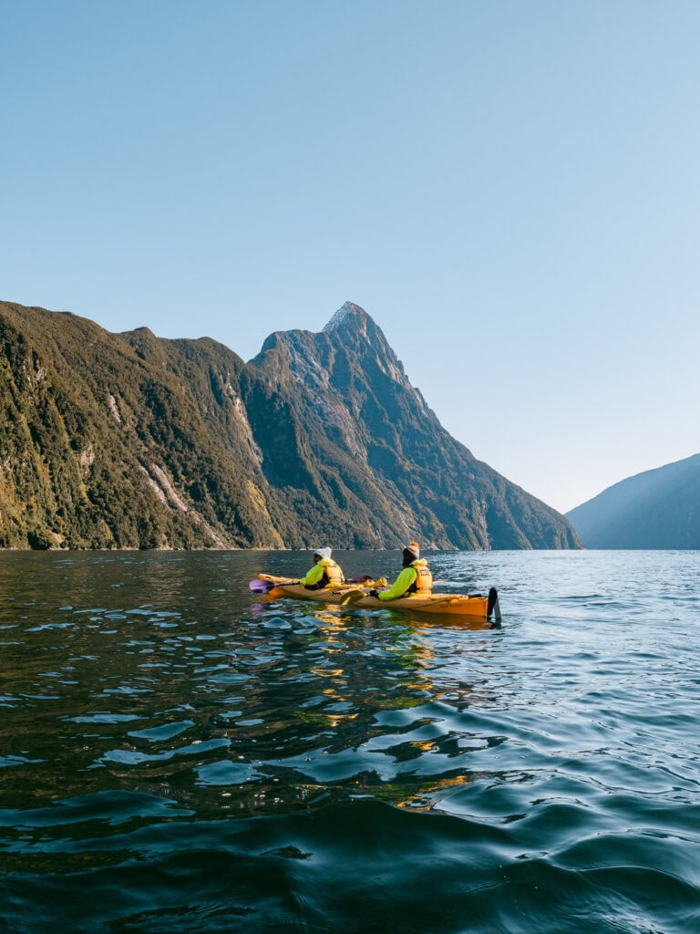 Two kayakers moving across the still surface of Milford Sound with jagged peaks in the distance