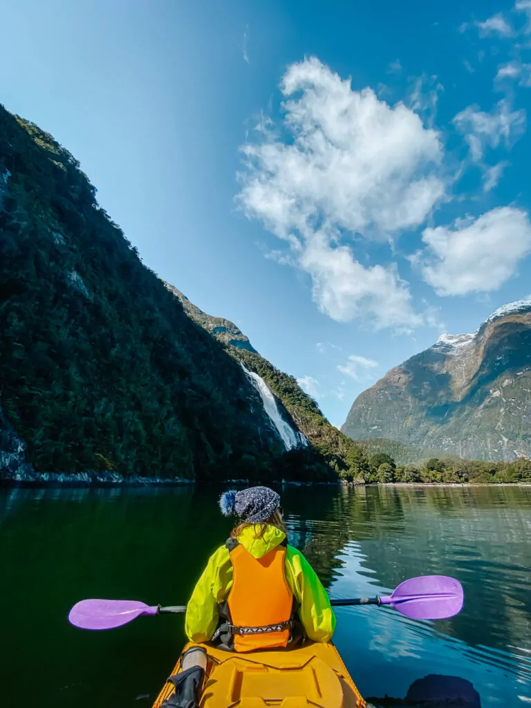 Person in a yellow jacket kayaking toward the towering cliffs of Milford Sound on a clear day