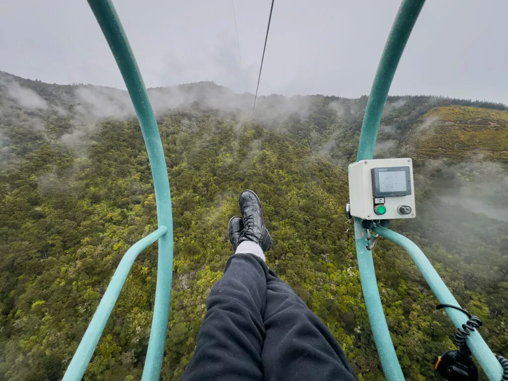 Person riding the Skywire zipline above lush forest canopy with legs stretched forward and misty hills ahead