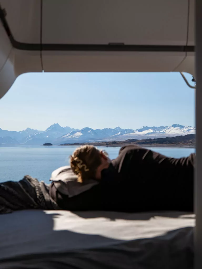 Traveller resting inside a campervan with wide window view of Lake Pukaki and Aoraki in the distance