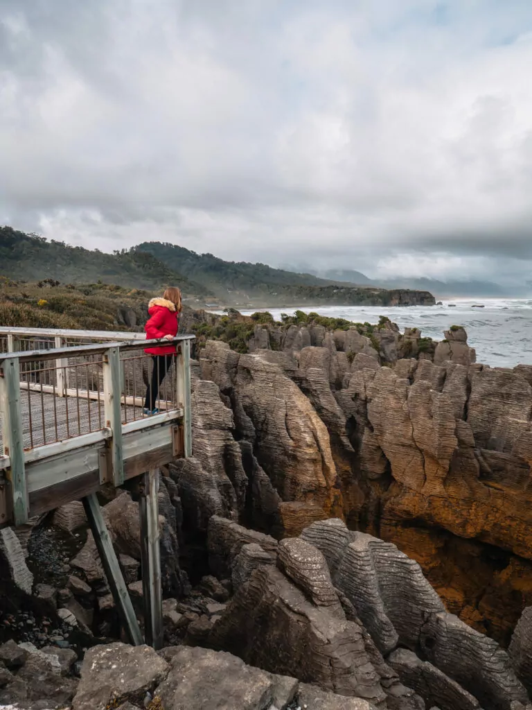 Person in red jacket standing on the lookout platform above crashing waves at Punakaiki’s Pancake Rocks and blowholes