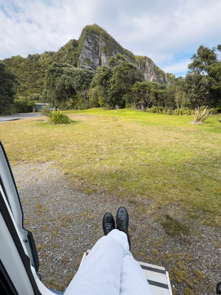 Feet hanging from campervan at Punakaiki Beach Camp with open grassy site, forest edge, and limestone cliffs beyond