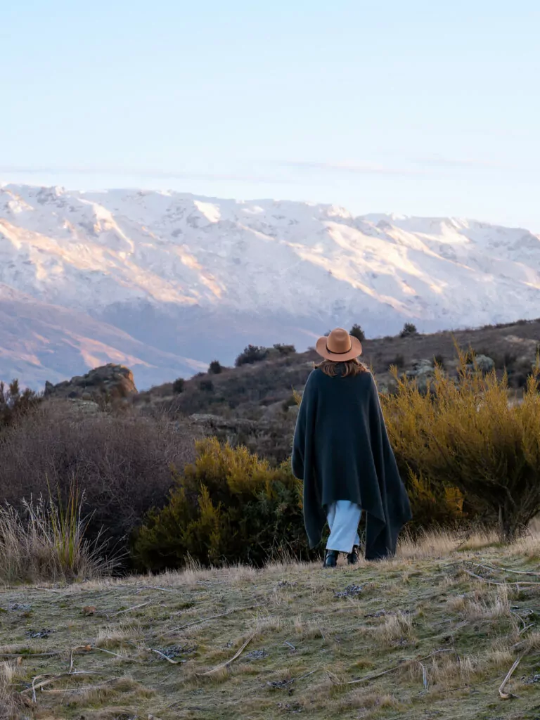 Woman in a blanket walking toward golden hills behind the Taima PurePod cabin near Cromwell at sunrise