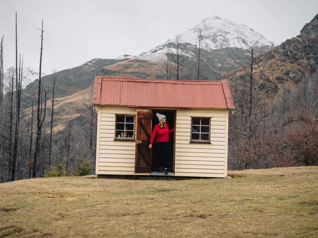 Small cream and red historic hut set in a rugged valley of Skippers Canyon with snow-capped peaks rising behind