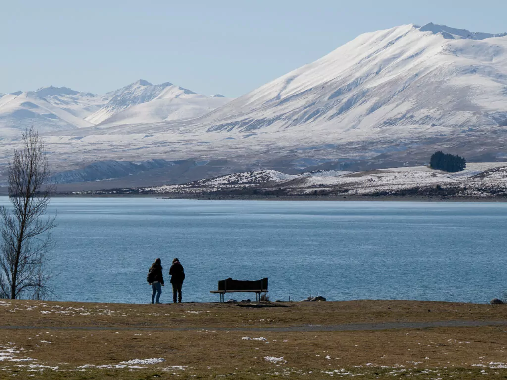 Silhouetted figures standing by the icy shoreline of Lake Tekapo with snow-dusted mountains beyond