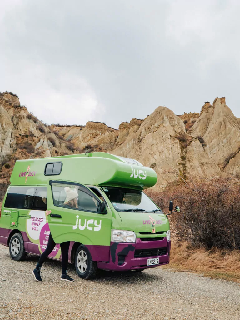JUCY campervan parked at the base of the towering, jagged formations of the Ōmarama Clay Cliffs