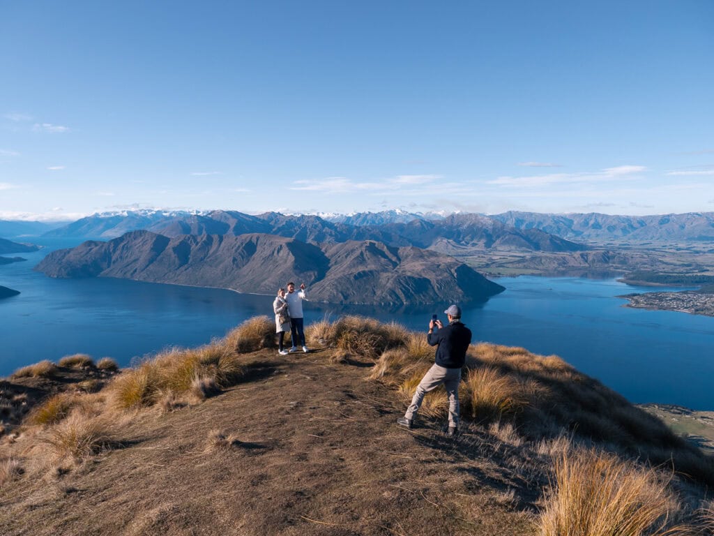 Two hikers walking along the grassy ridge of Coromandel Peak overlooking Lake Wānaka under clear blue skies
