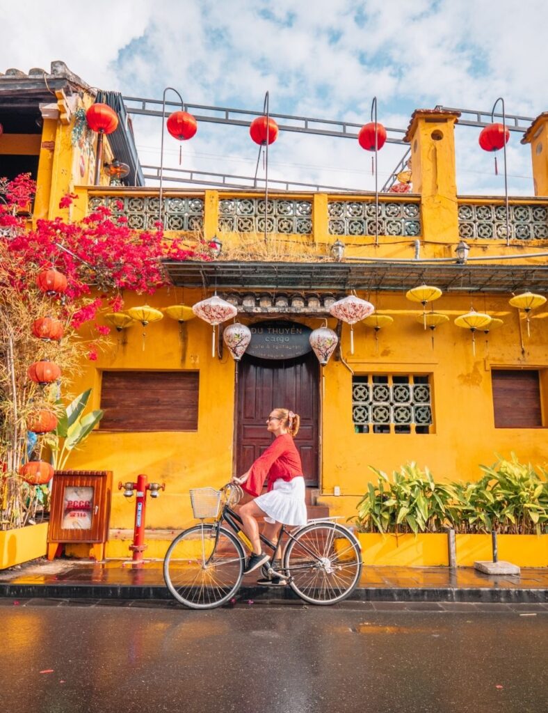 alexx riding a bike in hoi an, in front of a vibrant yellow building with hanging lanterns, a must-visit on any vietnam itinerary