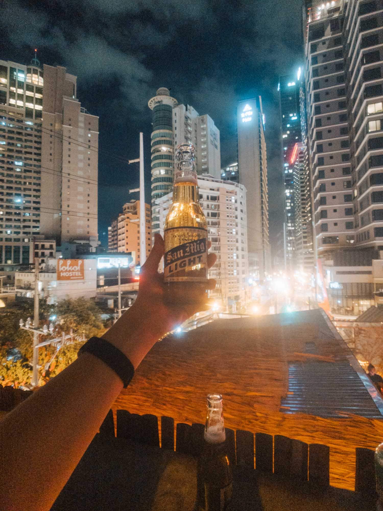 Holding a San Miguel Light on a Manila hostel rooftop with high-rises lit up at night