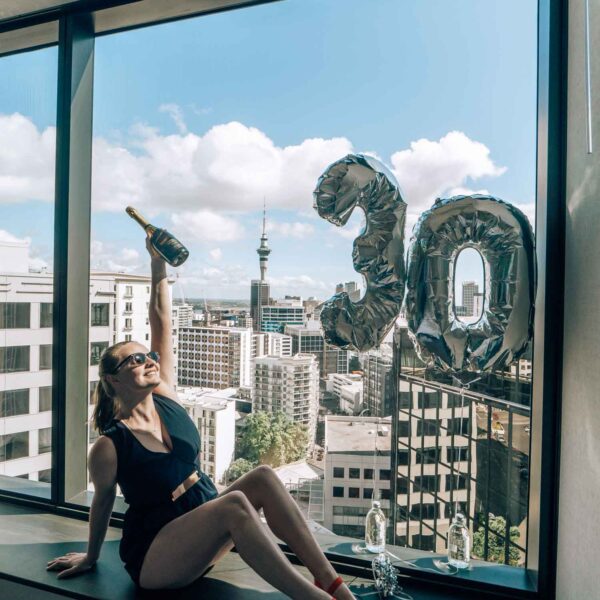 alexx sitting on window seat with auckland view holding a champagne bottle next to balloons that say 30