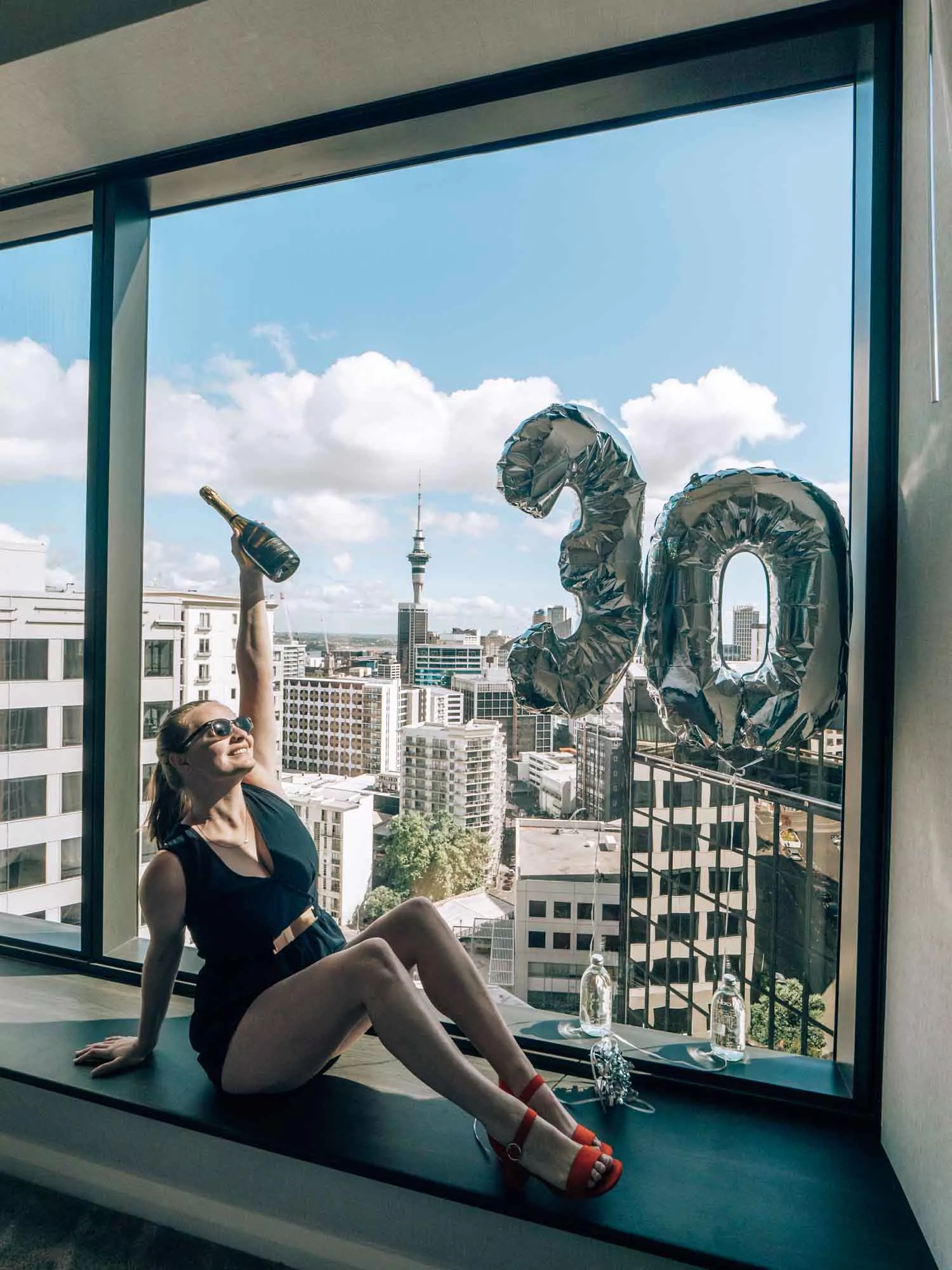 alexx sitting on window seat with auckland view holding a champagne bottle next to balloons that say 30