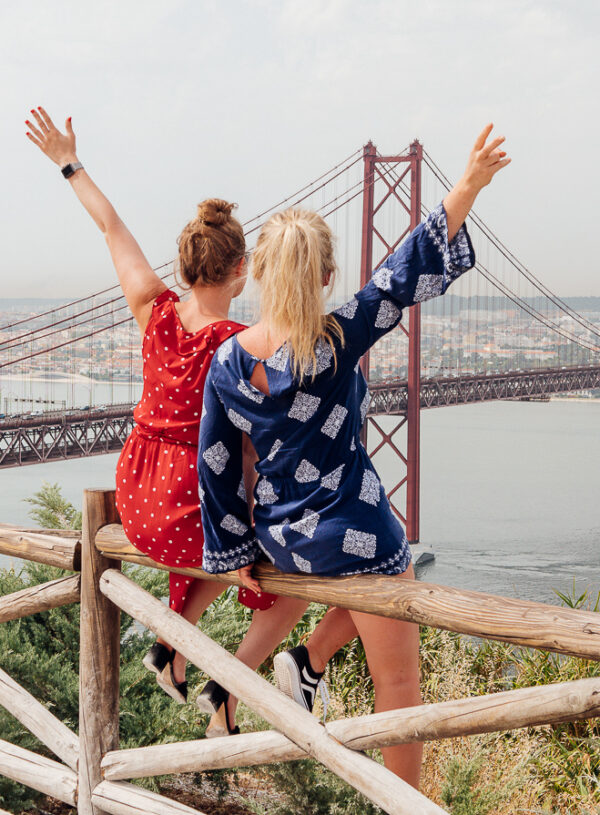 two girls sitting on a fence in front of lisbon bridge