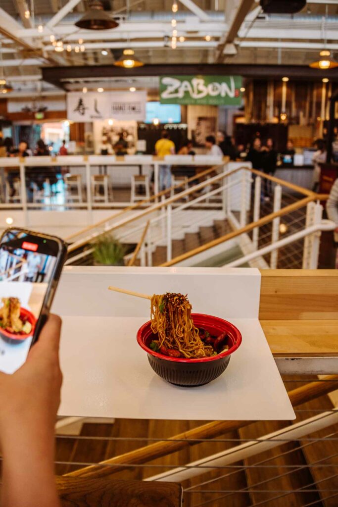 First-person perspective of a person photographing a bowl of ramen with floating chopsticks, in a food court with the sign 'ZABON' in the background